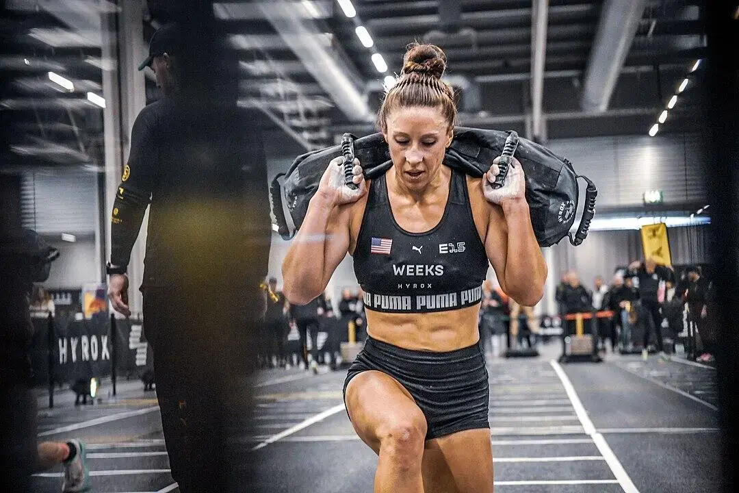 female athlete carrying a sandbag during a HYROX indoor fitness race.
